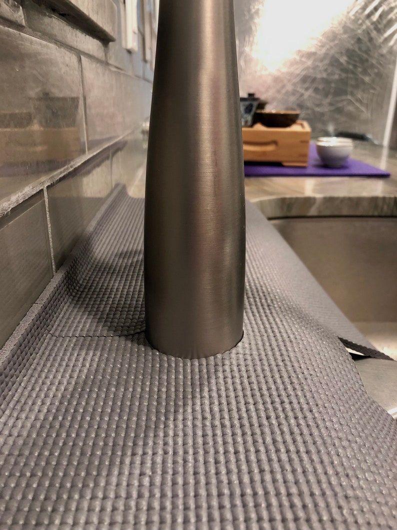May include: A gray yoga mat with a textured surface is positioned near a stainless steel faucet. The mat is rolled out on a countertop, with a sink visible in the background. The faucet is cylindrical and has a brushed metal finish.