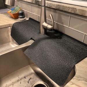 May include: A black sink mat with a textured surface, designed to fit around a faucet. The mat is placed over a stainless steel sink, with a faucet in the center. The mat is approximately 24 inches long and 12 inches wide.