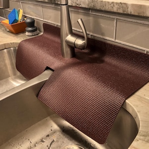 May include: A burgundy sink mat with a textured surface is placed around a stainless steel faucet. The mat is designed to protect the countertop from water damage. A stainless steel sink is in the foreground.