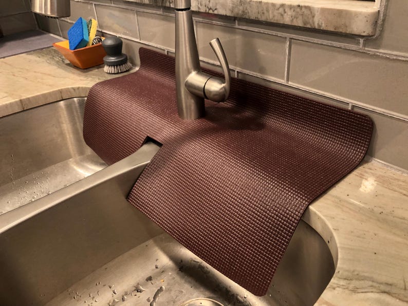 May include: A burgundy sink mat with a textured surface is placed around a stainless steel faucet. The mat is designed to protect the countertop from water damage. A stainless steel sink is in the foreground.