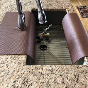 May include: A brown sink mat with a textured surface, designed to fit around a kitchen faucet. The mat is placed over a stainless steel sink, with cleaning brushes visible inside. The countertop is granite.