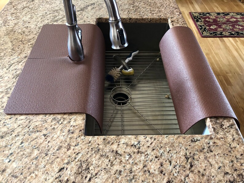 May include: A brown sink mat with a textured surface, designed to fit around a kitchen faucet. The mat is placed over a stainless steel sink, with cleaning brushes visible inside. The countertop is granite.