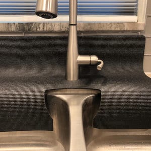 Op de afbeelding: Een zwarte, getextureerde gootsteenbeschermingsmat ligt achter een roestvrijstalen kraan. De mat heeft een gebogen ontwerp om rond de kraan en de gootsteen te passen. De mat is ontworpen om water op te vangen en het aanrecht te beschermen.
