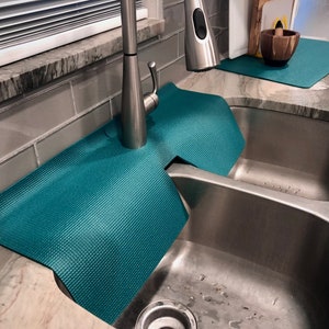 May include: Teal sink mats with a textured surface surround a stainless steel kitchen sink. The mats are designed to protect the worktop from water damage. A stainless steel tap is visible.