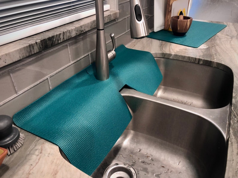 May include: Teal sink mats with a textured surface surround a stainless steel kitchen sink. The mats are designed to protect the worktop from water damage. A stainless steel tap is visible.