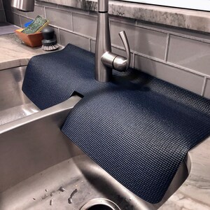May include: A navy blue sink mat with a textured surface, designed to fit around a faucet. The mat is placed in a stainless steel sink, offering protection against water stains and scratches. The mat's dimensions are approximately 24 inches long and 14 inches wide.