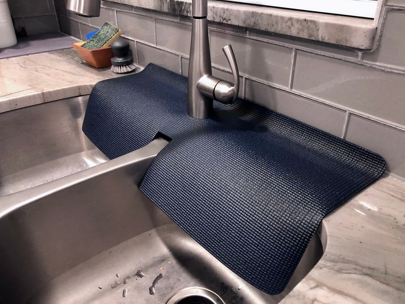 May include: A navy blue sink mat with a textured surface, designed to fit around a faucet. The mat is placed in a stainless steel sink, offering protection against water stains and scratches. The mat's dimensions are approximately 24 inches long and 14 inches wide.