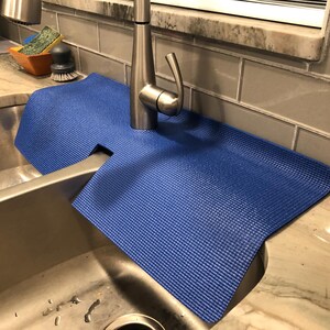 May include: A blue sink mat with a textured surface is placed around a stainless steel faucet. The mat is designed to catch water and protect the countertop. A stainless steel sink is in the foreground.