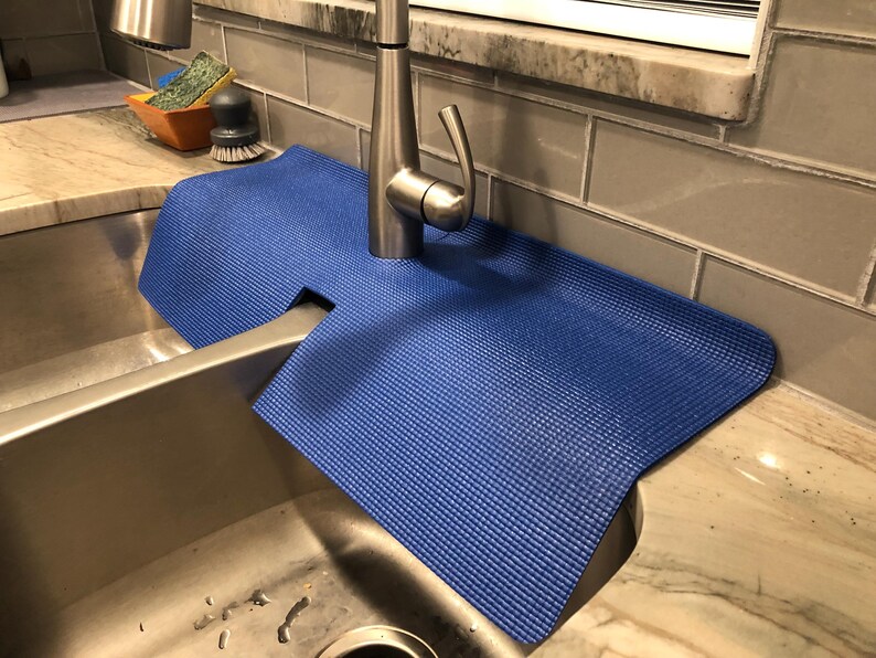 May include: A blue sink mat with a textured surface is placed around a stainless steel faucet. The mat is designed to catch water and protect the countertop. A stainless steel sink is in the foreground.