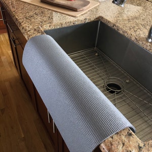 May include: A gray, textured sink mat is draped over the edge of a granite countertop. The mat is rolled up, revealing its textured surface. A stainless steel sink and a cutting board with apples and a knife are visible in the background.