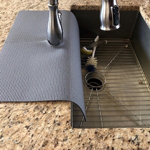 May include: A gray sink mat with a textured surface partially covers a stainless steel sink. A stainless steel faucet is visible, along with cleaning brushes and a metal sink rack. The countertop is granite.