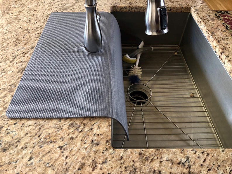 May include: A gray sink mat with a textured surface partially covers a stainless steel sink. A stainless steel faucet is visible, along with cleaning brushes and a metal sink rack. The countertop is granite.