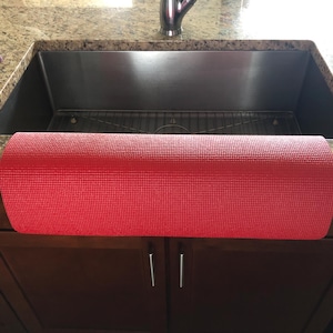 May include: A rolled-up red yoga mat is positioned in front of a stainless steel kitchen sink. The mat is made of a textured material. The sink is set into a granite countertop, with dark brown cabinets below.