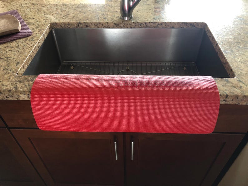 May include: A rolled-up red yoga mat is positioned in front of a stainless steel kitchen sink. The mat is made of a textured material. The sink is set into a granite countertop, with dark brown cabinets below.
