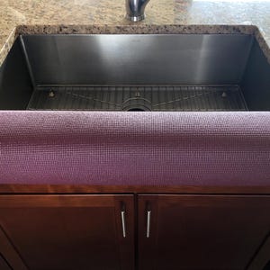May include: A rolled-up, textured, purple yoga mat rests on a dark brown cabinet in front of a stainless steel kitchen sink. The sink is set into a granite countertop.