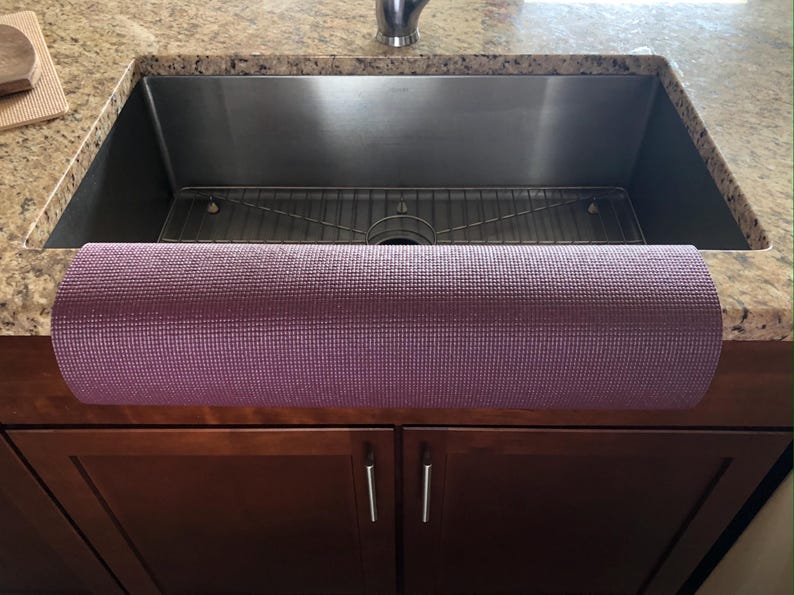 May include: A rolled-up, textured, purple yoga mat rests on a dark brown cabinet in front of a stainless steel kitchen sink. The sink is set into a granite countertop.