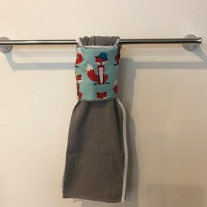 May include: A gray hand towel with a white border hangs from a chrome towel bar. The top portion features a light blue fabric with a repeating pattern of red foxes wearing hats. The towel is made of a textured material.