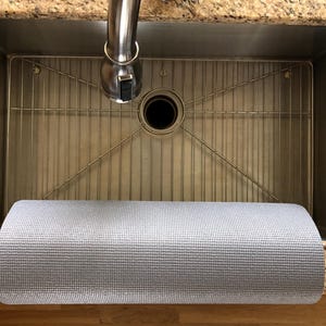 May include: A stainless steel kitchen sink with a metal grid and a rolled-up gray mat. The sink has a faucet and is set into a granite countertop. The mat is made of a textured material.
