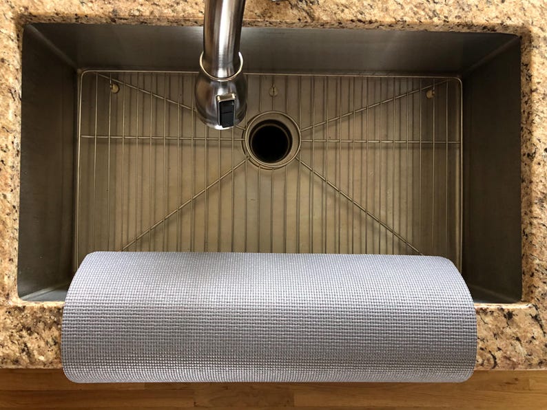 May include: A stainless steel kitchen sink with a metal grid and a rolled-up gray mat. The sink has a faucet and is set into a granite countertop. The mat is made of a textured material.