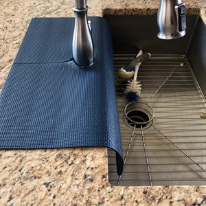 May include: A dark blue sink mat made of a textured material, partially covering a stainless steel sink. The mat is positioned next to a brushed nickel faucet. Cleaning brushes and a sink strainer are visible inside the sink.