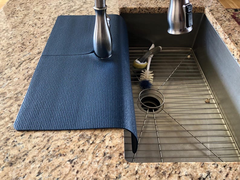 May include: A dark blue sink mat made of a textured material, partially covering a stainless steel sink. The mat is positioned next to a brushed nickel faucet. Cleaning brushes and a sink strainer are visible inside the sink.