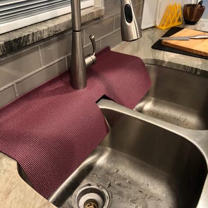 May include: A burgundy sink mat is draped over a stainless steel double sink. The mat is made of a textured material and is designed to protect the countertop from water damage. A stainless steel faucet is visible in the background.