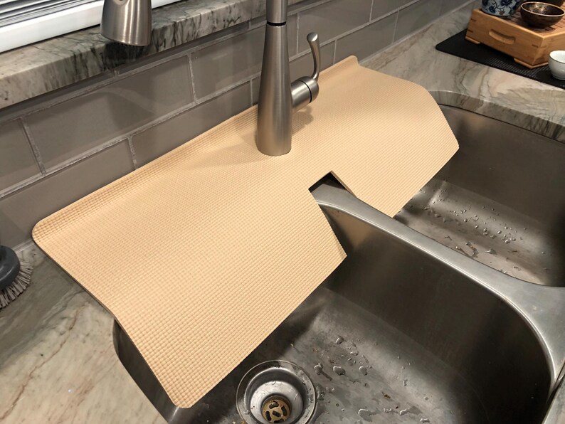 May include: A beige sink mat with a textured surface, designed to fit around a faucet. The mat is placed over a stainless steel sink, offering protection and a clean look. The faucet is made of brushed metal.