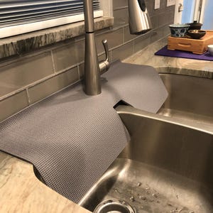 May include: A gray sink mat with a textured surface is draped over a stainless steel double sink. The mat is positioned around a silver faucet. A small tea set with a wooden tray and purple mat is on the countertop.