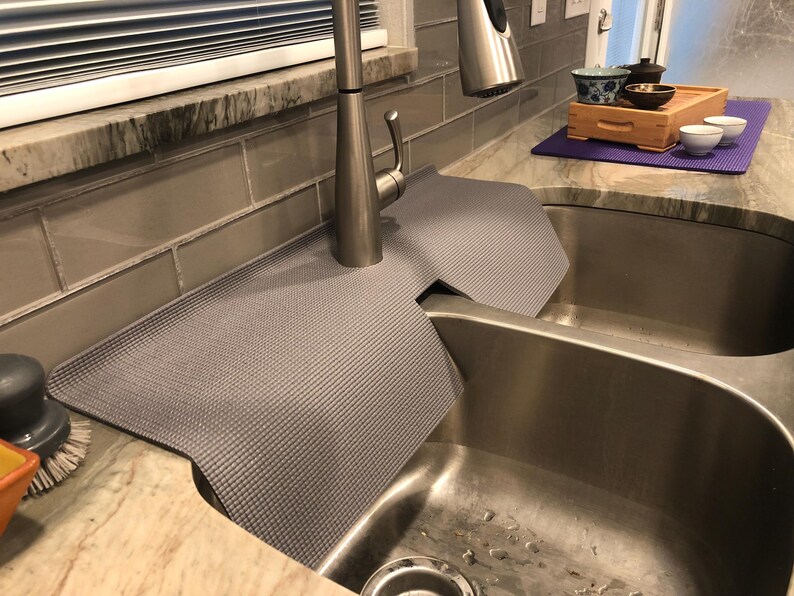 May include: A gray sink mat with a textured surface is draped over a stainless steel double sink. The mat is positioned around a silver faucet. A small tea set with a wooden tray and purple mat is on the countertop.