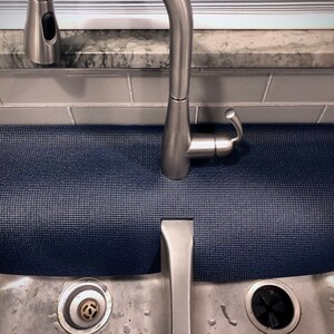May include: A navy blue sink mat with a textured surface, positioned beneath a stainless steel faucet. The mat is rectangular and designed to protect the sink area from water and scratches. The sink is made of stainless steel.