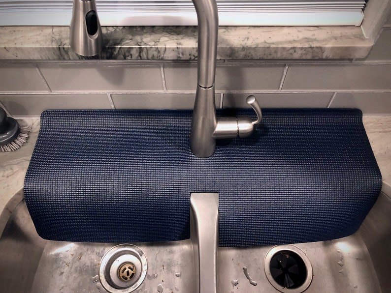 May include: A navy blue sink mat with a textured surface, positioned beneath a stainless steel faucet. The mat is rectangular and designed to protect the sink area from water and scratches. The sink is made of stainless steel.