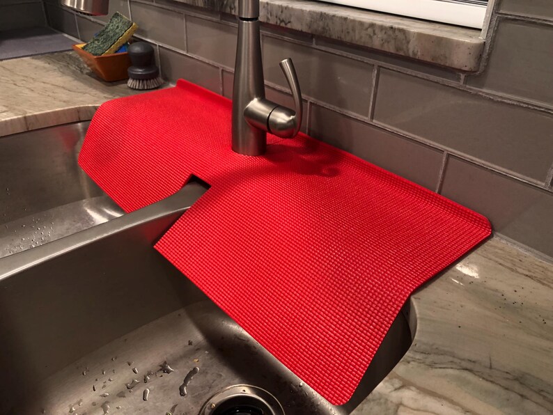 May include: A bright red, textured sink mat designed to fit around a faucet. The mat is made of a flexible material and sits on the countertop and inside the stainless steel sink. The mat is designed to catch water and protect the countertop.