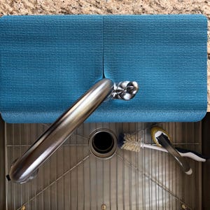 May include: A teal blue sink mat rests on a granite countertop, partially covering a stainless steel sink. A chrome faucet extends over the sink, with a brush and a sponge resting inside. The mat is textured and rectangular.