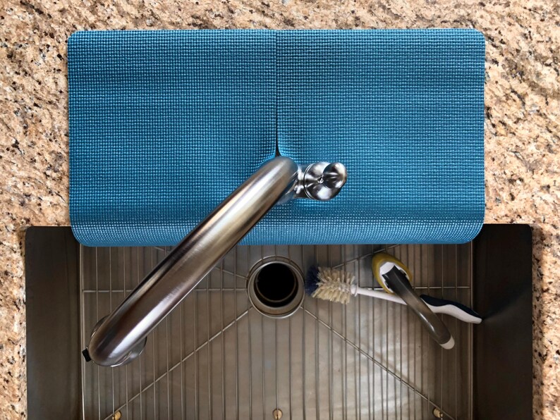 May include: A teal blue sink mat rests on a granite countertop, partially covering a stainless steel sink. A chrome faucet extends over the sink, with a brush and a sponge resting inside. The mat is textured and rectangular.