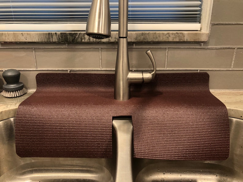 Puede incluir: Una estera de fregadero de goma marr&oacute;n con una superficie texturizada, dise&ntilde;ada para ajustarse alrededor de un grifo de fregadero de cocina. La estera se coloca en un fregadero de acero inoxidable.