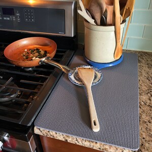 May include: A copper-colored frying pan with food inside sits on a black stovetop. A gray mat protects the countertop, with a wooden spatula and a ceramic crock filled with wooden cooking utensils. A blue and white plate is also on the counter.