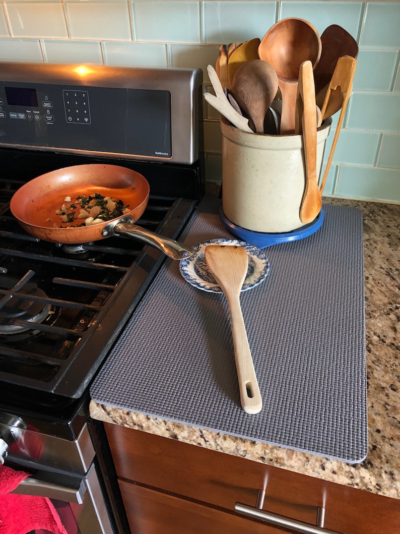 May include: A copper-colored frying pan with food inside sits on a black stovetop. A gray mat protects the countertop, with a wooden spatula and a ceramic crock filled with wooden cooking utensils. A blue and white plate is also on the counter.