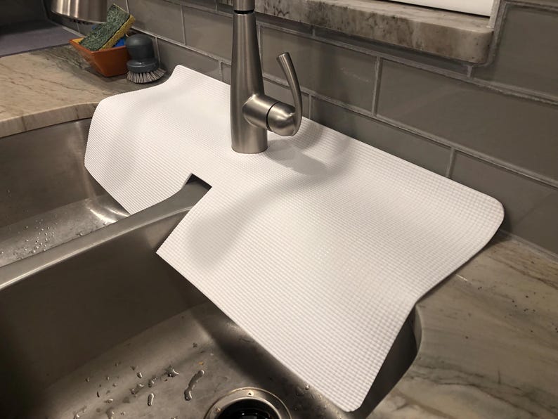 May include: A white, textured sink mat is placed over a stainless steel sink. The mat has a cutout for the faucet and is designed to protect the sink's surface. The faucet is made of brushed nickel.