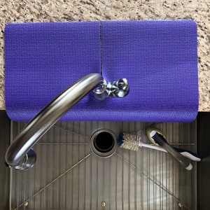 May include: Overhead view of a stainless steel kitchen sink with a chrome faucet. A rolled-up purple mat rests on the counter. A brush and sponge are in the sink.