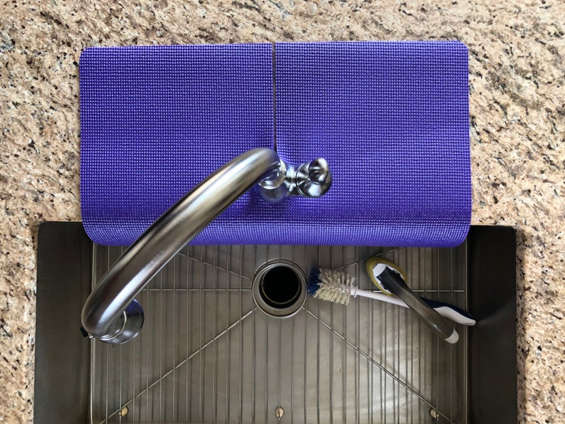 May include: Overhead view of a stainless steel kitchen sink with a chrome faucet. A rolled-up purple mat rests on the counter. A brush and sponge are in the sink.