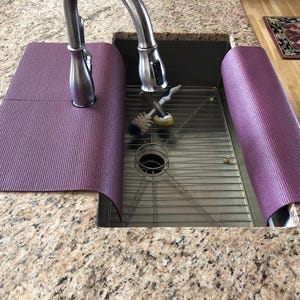 May include: A purple sink mat is wrapped around the faucet and the sink's edge. The mat is made of a textured material and is designed to protect the countertop from water damage. The sink is stainless steel and has a metal grate.