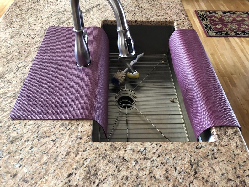 May include: A purple sink mat is wrapped around the faucet and the sink's edge. The mat is made of a textured material and is designed to protect the countertop from water damage. The sink is stainless steel and has a metal grate.