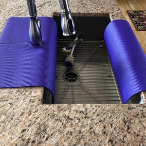 May include: A stainless steel kitchen sink with a metal grate and a faucet. Two rolled-up, textured, purple mats are placed on the countertop on either side of the sink. Cleaning brushes are inside the sink.
