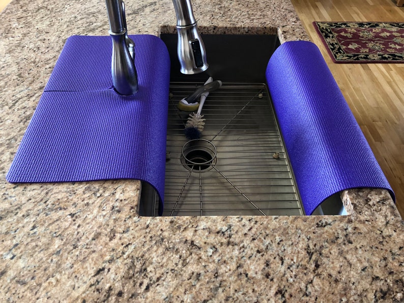May include: A stainless steel kitchen sink with a metal grate and a faucet. Two rolled-up, textured, purple mats are placed on the countertop on either side of the sink. Cleaning brushes are inside the sink.