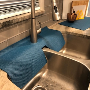 May include: A blue sink mat is placed around a stainless steel faucet and sink. The mat is made of a textured material and is designed to protect the countertop from water damage. Additional kitchen accessories are visible in the background.