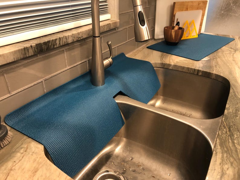 May include: A blue sink mat is placed around a stainless steel faucet and sink. The mat is made of a textured material and is designed to protect the countertop from water damage. Additional kitchen accessories are visible in the background.