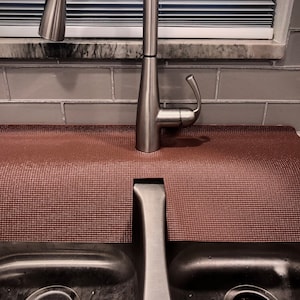 May include: A brown sink mat with a textured surface is placed under a stainless steel faucet. The mat is designed to protect the countertop from water damage. The sink is stainless steel.