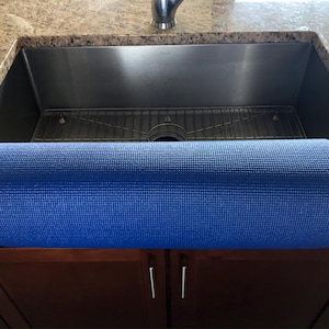 May include: A rolled-up blue yoga mat rests on a dark brown cabinet in front of a stainless steel sink. The sink is set into a granite countertop. The mat is a solid, textured blue.
