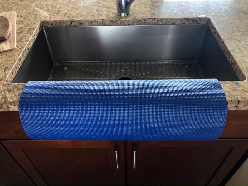 May include: A rolled-up blue yoga mat rests on a dark brown cabinet in front of a stainless steel sink. The sink is set into a granite countertop. The mat is a solid, textured blue.