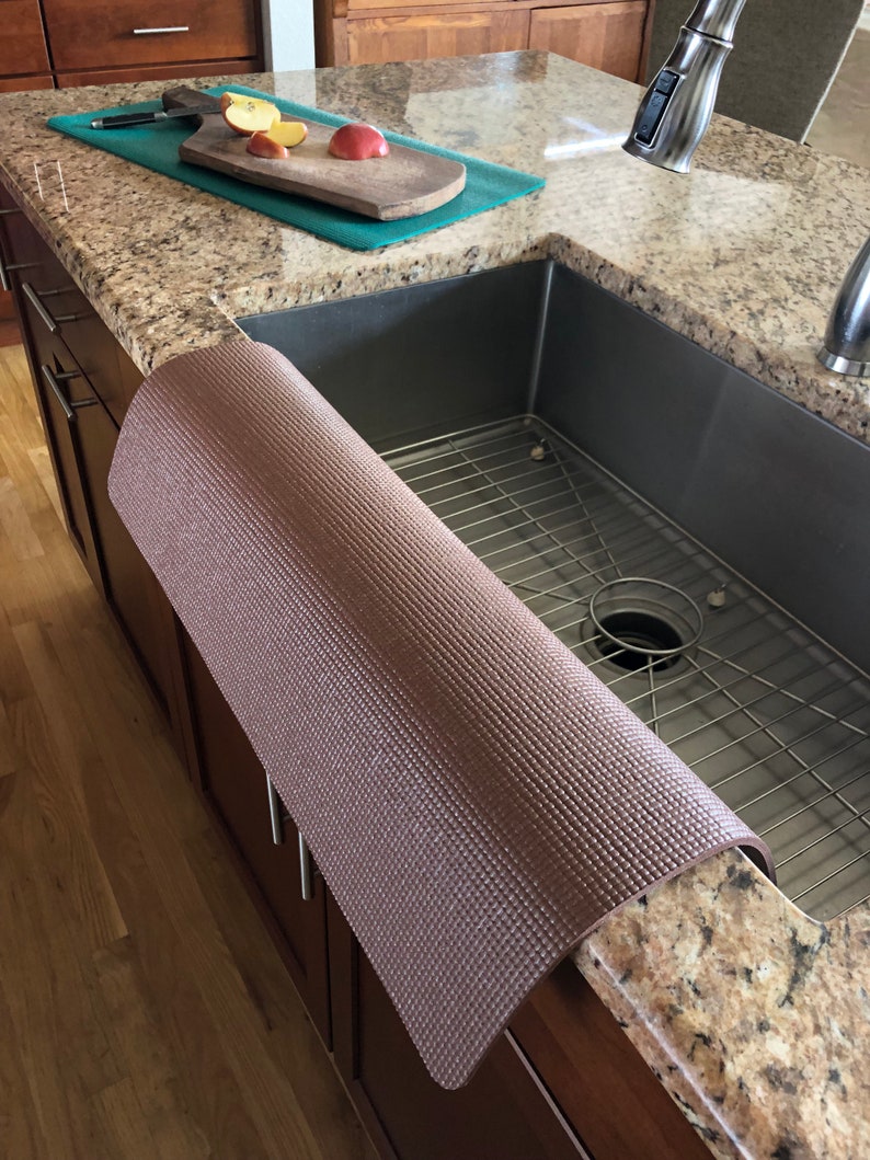 May include: A rolled-up brown sink protector mat with a textured surface rests on a granite countertop next to a stainless steel sink. A cutting board with sliced apples sits on a teal mat.